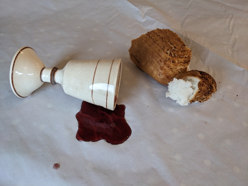 Spilt wine from pottery chalice and broken bread all on white cloth